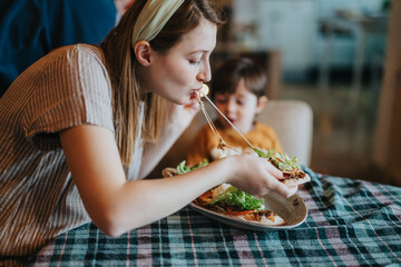 A woman, sitting at a table, eats a meal while a young child watches. The scene conveys warmth and...