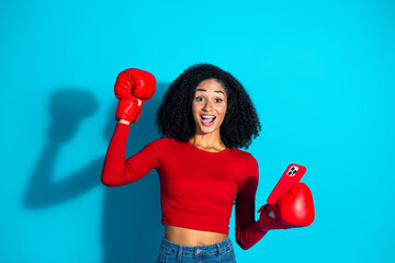 Young woman in red boxing gloves holding a smartphone with a cheerful expression on a vibrant blue...