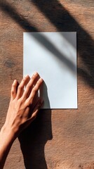 Hand Placing Blank White Sheet of Paper on Rustic Wooden Table with Soft Shadows and Warm Lighting