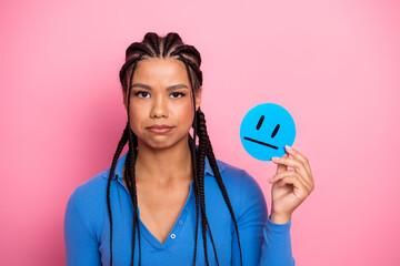 Charming young woman holding a blue sad face on a pink background, wearing a stylish blue shirt with braided hair