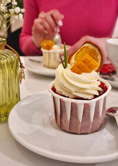 Closeup of a cupcake dessert topped with whipped cream, a slice of dried orange, a piece of caramel. Young woman in the background tasting the confectionery