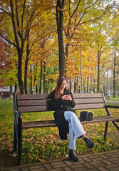 Aesthetic young woman wearing a black coat sitting on a wooden bench in the autumn park, using her phone. Female relaxing surrounded by trees with colorful foliage