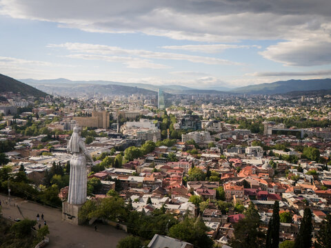 Aerial view of Kartlis Deda statue overlooking the city's terracotta rooftops and modern skyline under a sky of dramatic clouds, Tbilisi, Tbilisi, Georgia.