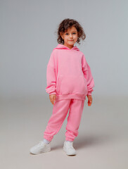Child stands with one hand on her hip, wearing a vibrant pink tracksuit and white sneakers. She displays a playful expression against a simple backdrop.