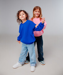 Two young girls stand confidently, showcasing colorful sweatshirts and relaxed jeans in a cheerful studio setting. Their playful expressions highlight their friendship and style.