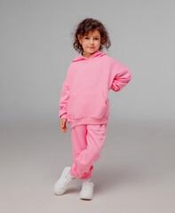 A child stands with one hand on her hip, wearing a vibrant pink tracksuit and white sneakers. She displays a playful expression against a simple backdrop.