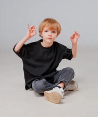 A young child sits on the floor cross-legged, wearing a black oversized shirt and jeans. The child makes hand gestures, displaying a playful and relaxed demeanor in a simple studio.