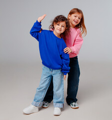 Two young girls stand in a studio, wearing vibrant sweatshirts and denim jeans. They smile and strike playful poses, expressing happiness and friendship in a fun atmosphere.