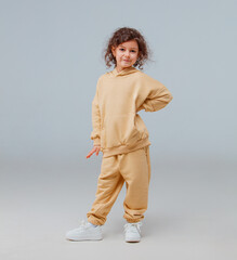 A young girl stands with her hand on her hip, showcasing her beige clothing in a bright studio space. Her curly hair adds to her playful demeanor as she strikes a fun pose.