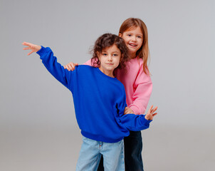 Two girls stand close together, wearing colorful sweaters, with one extending her arm in a playful gesture. Their joyful expressions highlight the fun of their indoor playtime.