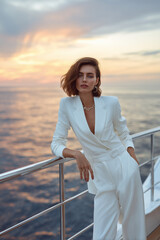 Fototapeta premium Elegant woman in stylish white suit posing on yacht at sunset with serene ocean view in the background