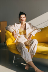 Confident young woman in stylish white suit and yellow top sitting barefoot on a modern yellow sofa in natural sunlight