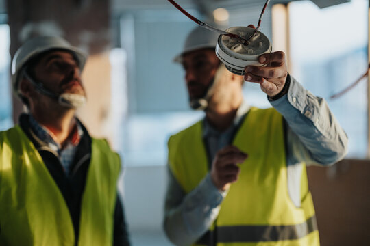 Focused engineers wearing safety gear examine a smoke detector at a construction area, ensuring proper compliance with installation standards and safety protocols.