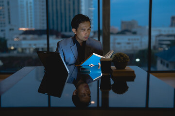 Asian businessman reading a book while sitting at his desk in a modern office at night, with city lights in the background and a tablet next to him