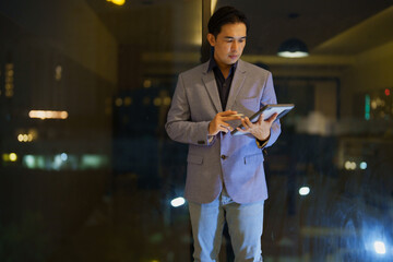 Focused business professional stands by a window, using a tablet. The city lights create a modern,...