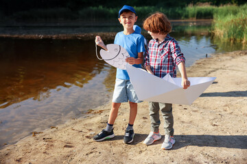 Cute little boys with paper boat and fish by lake in nature