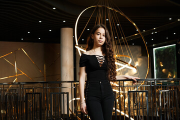 A girl in black pants near a metal railing with a large chandelier behind her