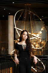 A girl in black pants near a metal railing with a large chandelier behind her