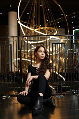 A girl in black pants near a metal railing with a large chandelier behind her. She is sitting on the ground