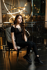 A girl in black trousers sits on a chair near a metal railing, behind which hangs a large chandelier.