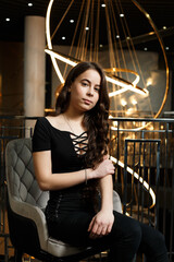 A girl in black trousers sits on a chair near a metal railing, behind which hangs a large chandelier.
