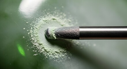 Close-up view of a metallic tool interacting with pale green powder, creating a textured surface. The powder is scattered on a dark green background.