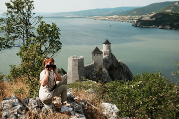 Woman sitting on rocks using binoculars to explore the view of Golubac Fortress and the Danube River. Travel in Serbia concept