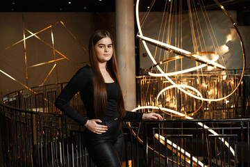 A girl at a metal railing with a large chandelier hanging behind it. She is wearing leather pants and a black blouse.