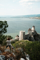 Woman standing on hillside looking at medieval Golubac Fortress on the Danube River. Rear view....