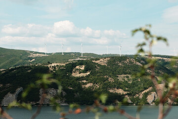 Wind turbines on green hills above River. Renewable energy landscape on a partly cloudy summer day,...