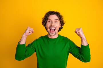 Excited young man in a vibrant green sweater celebrating with joyful gestures against a yellow...