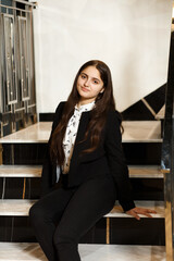 Girl in black pants on the stairs indoors, she is wearing a white blouse and jacket, she is sitting on the steps