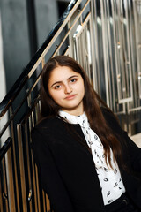 Girl in black pants on the stairs indoors, she is wearing a white blouse and jacket, she is sitting on the steps