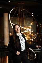 A girl at a metal railing with a large chandelier hanging behind it. She is wearing black trousers and a white blouse with a jacket.