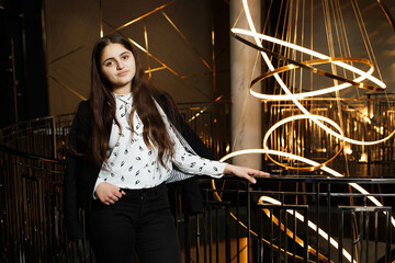 A girl at a metal railing with a large chandelier hanging behind it. She is wearing black trousers and a white blouse with a jacket.