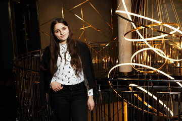 A girl at a metal railing with a large chandelier hanging behind it. She is wearing black trousers and a white blouse with a jacket.