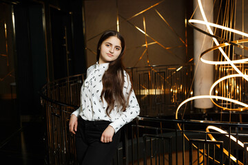 A girl at a metal railing with a large chandelier hanging behind it. She is wearing black trousers and a white blouse with a jacket.