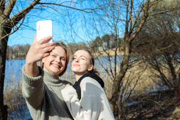 Two women have fun taking photos on a smartphone on a spring day in a park by the river. A beautiful woman and her mother have fun together.