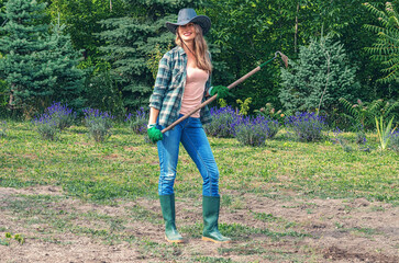 Satisfied woman in hat and rubber boots standing behind blooming lavender bushes and holding hoe.