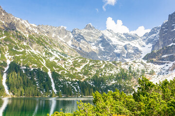 alpine lake in spring on a sunny day. travel landscape of rysy ridge in high tatra mountains of poland. snow among rocks and spruce forest on the hillside reflection in water. blue sky with clouds