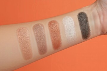 Close-up of shimmery eyeshadow swatches on an arm, displaying a cosmetic palette with nude, brown, and dark shades against an orange background.