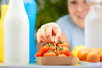 Smiling woman taking fresh tomatoes from inside a refrigerator. View from fridge with eggs, milk, and drinks in foreground. Healthy food and lifestyle concept.