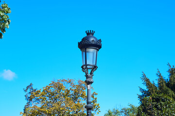 Decorative iron street lamp with crown details against clear blue sky and autumn trees