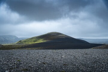 View of a solitary, dark volcanic cone rises starkly from a vast, grey gravel plain under a brooding sky, a scene of stark beauty, Kirkjubaejarklaustur, SkaftÃ¡rhreppur, Iceland.