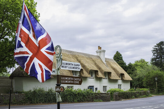 View of a thatched roof cottage behind a Union Jack flag on a signpost, with the sky reflecting off the road's surface, Wareham, England, United Kingdom.
