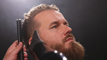 Young Man with Beard at Barber Shop. Barber is Preparing his Haircut. Dark Atmosphere, Black Background. Concept of Barber and a Client.