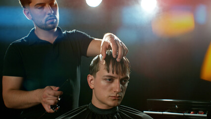 Young Man with Beard at Barber Shop. Barber is Preparing his Haircut. Dark Atmosphere, Black Background. Concept of Barber and a Client.
