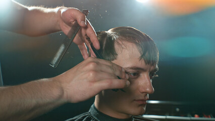 Young Man with Beard at Barber Shop. Barber is Preparing his Haircut. Dark Atmosphere, Black Background. Concept of Barber and a Client.