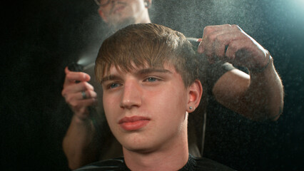 Young Man with Beard at Barber Shop. Barber is Preparing his Haircut. Dark Atmosphere, Black Background. Concept of Barber and a Client.