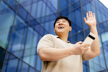 Asian businessman waving and holding smartphone in front of modern office building